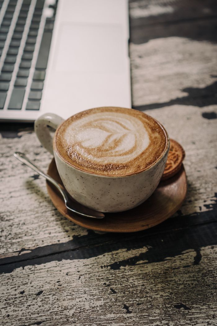 A close-up of a cappuccino cup and saucer on a rustic table next to a laptop.