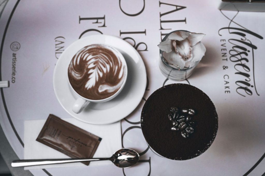 A stylish flatlay featuring intricate latte art and a chocolate dessert in a modern cafe setting.